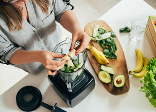 Top-down view of a woman breaking a banana in half so she can add it to the blender that is already partly full of smoothie ingredients.