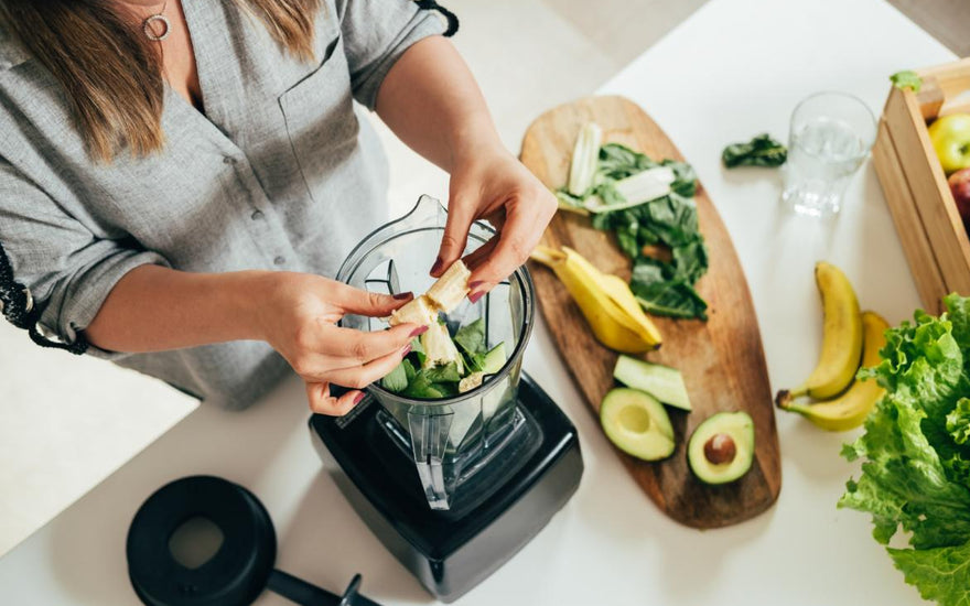 Top-down view of a woman breaking a banana in half so she can add it to the blender that is already partly full of smoothie ingredients.