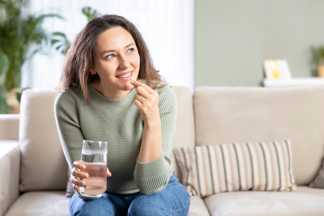 Woman in jeans and a green sweater sitting on a tan couch holding a glass of water while she puts a white pill in her mouth.
