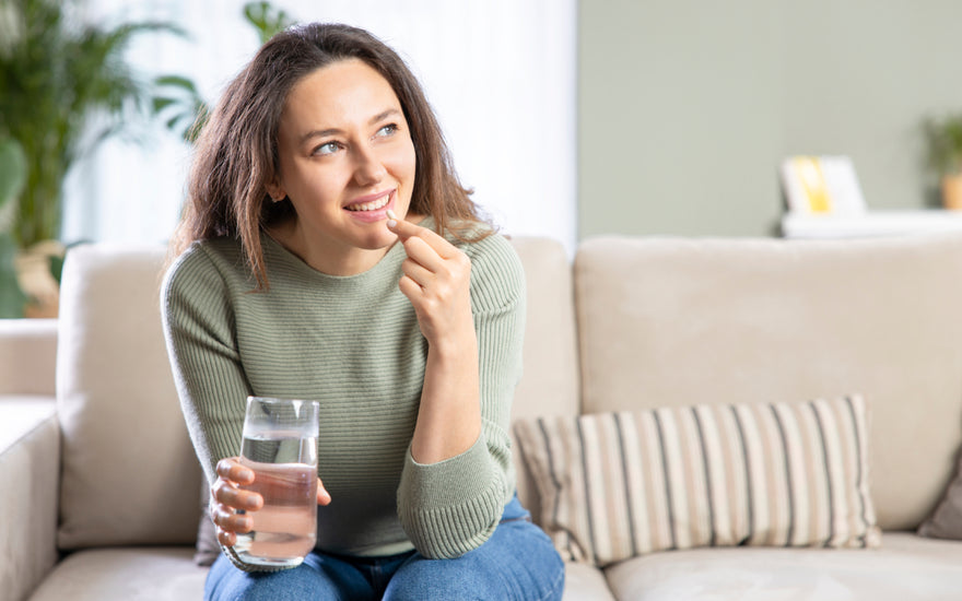 Woman in jeans and a green sweater sitting on a tan couch holding a glass of water while she puts a white pill in her mouth.