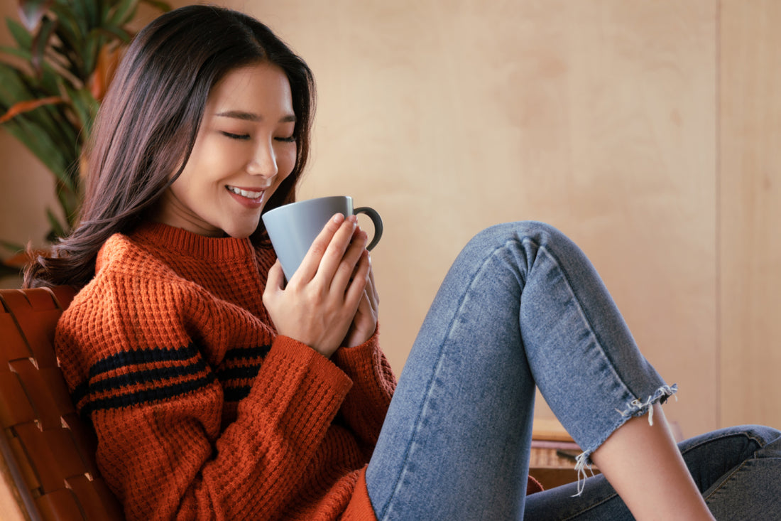 Young woman in jeans and an orange sweater sitting in a chair holding a coffee mug.