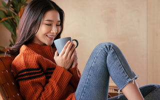 Young woman in jeans and an orange sweater sitting in a chair holding a coffee mug.