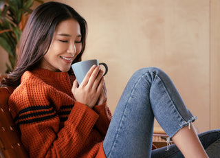 Young woman in jeans and an orange sweater sitting in a chair holding a coffee mug.