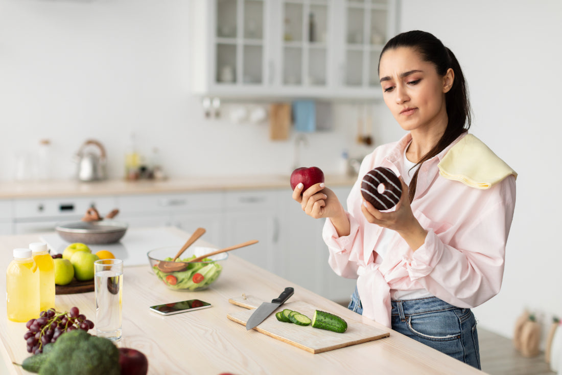 Woman in a pink blouse standing at the kitchen counter trying to decide between an apple and a donut.