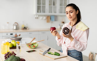 Woman in a pink blouse standing at the kitchen counter trying to decide between an apple and a donut.