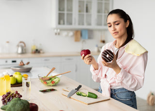 Woman in a pink blouse standing at the kitchen counter trying to decide between an apple and a donut.