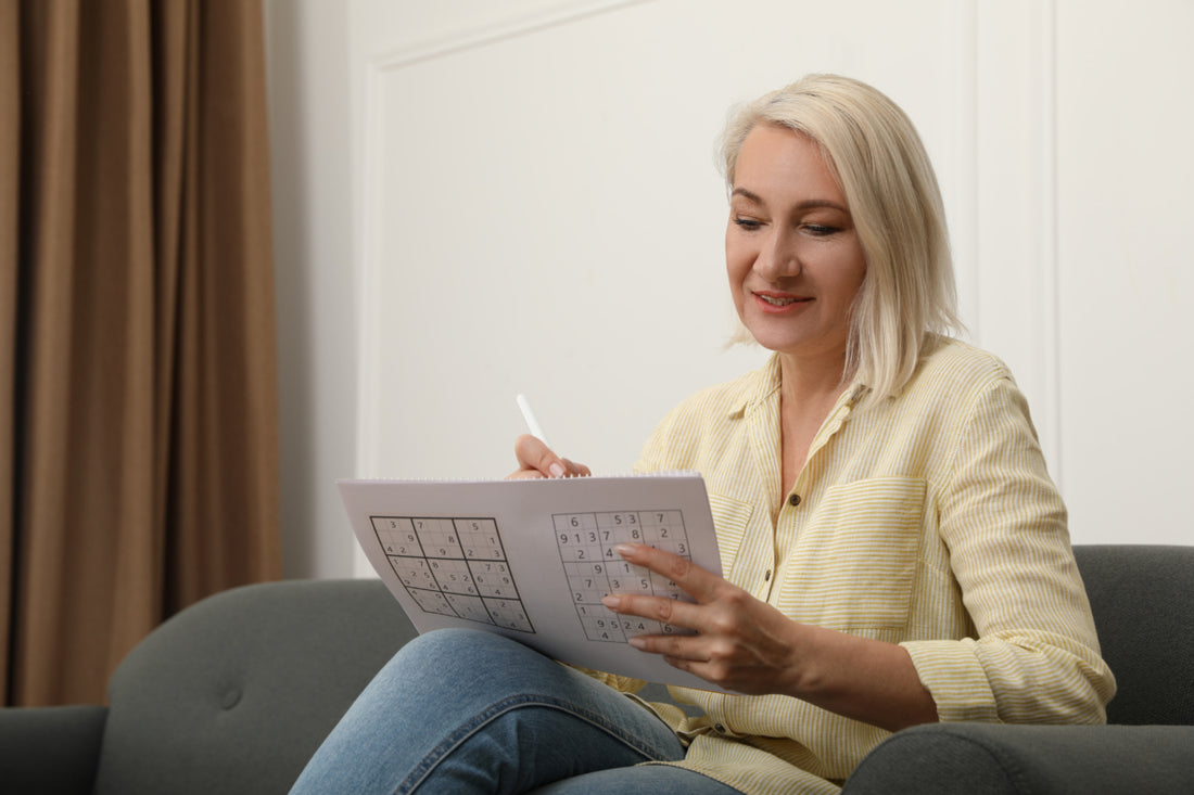 Middle ages woman in a yellow blouse sitting on a grey couch while she does a sudoku puzzle.