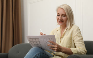 Middle ages woman in a yellow blouse sitting on a grey couch while she does a sudoku puzzle.