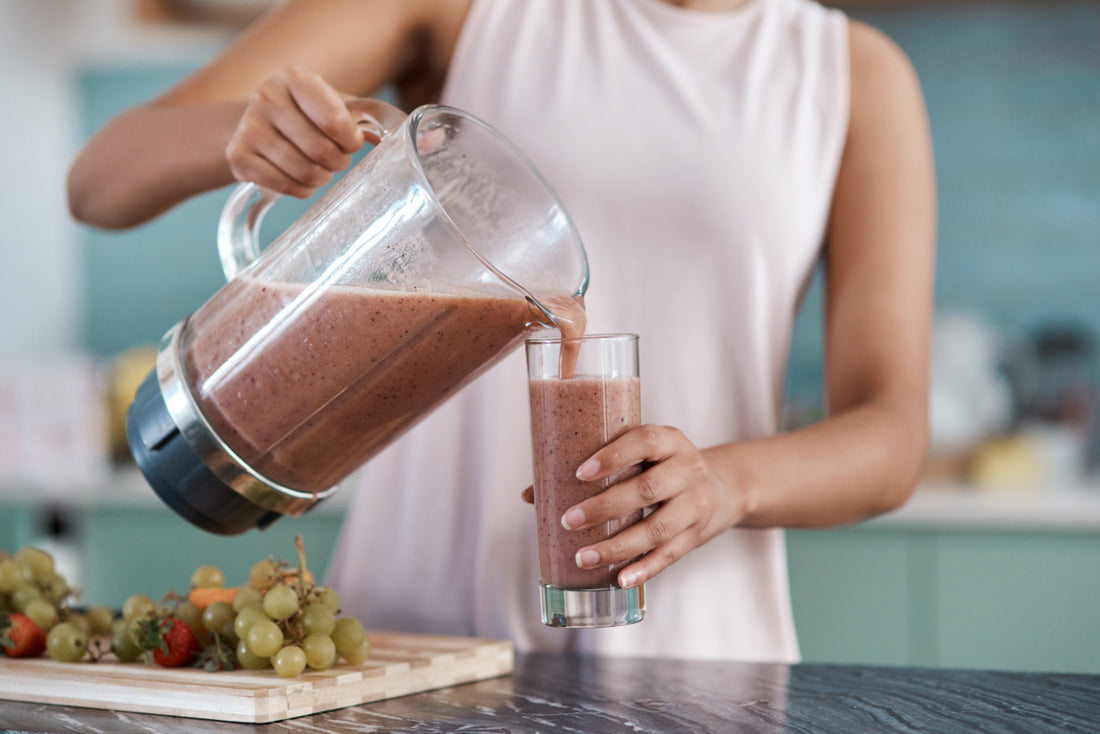 Woman in a pink tank top standing at a kitchen counter pouring a purple-colored smoothie our of a blender into a tall glass.
