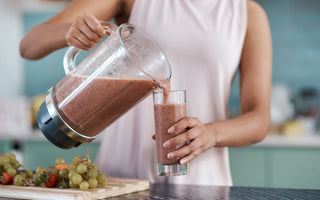 Woman in a pink tank top standing at a kitchen counter pouring a purple-colored smoothie our of a blender into a tall glass.