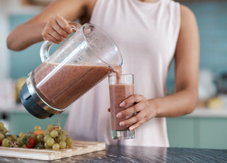 Woman in a pink tank top standing at a kitchen counter pouring a purple-colored smoothie our of a blender into a tall glass.