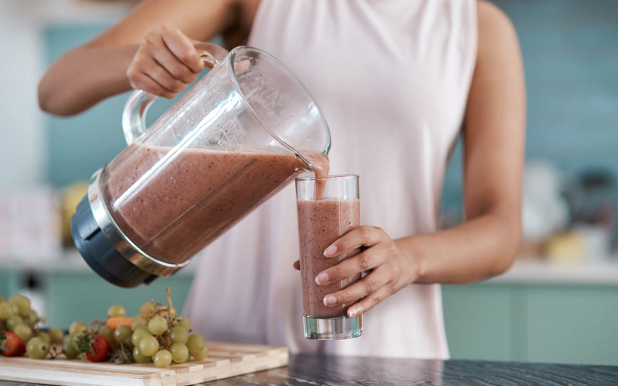 Woman in a pink tank top standing at a kitchen counter pouring a purple-colored smoothie our of a blender into a tall glass.