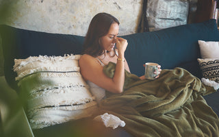 Woman sitting on a blue couch covered in a green blanket. She has a blue mug in one hand and is pinching the bridge of her nose with the fingers on the other hand.