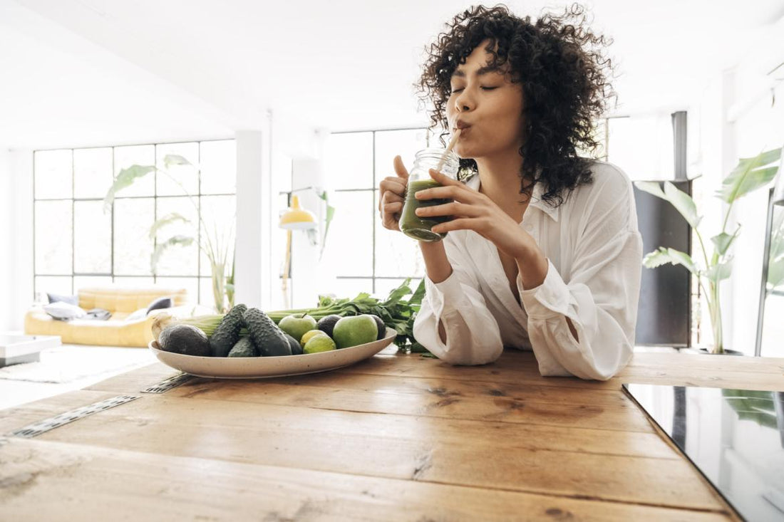 Woman in a white button up shirt sitting at a wooden kitchen table sipping a green smoothie out of a glass mug.