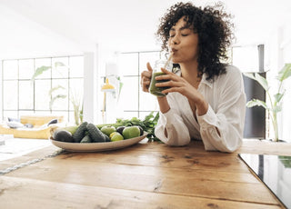 Woman in a white button up shirt sitting at a wooden kitchen table sipping a green smoothie out of a glass mug.