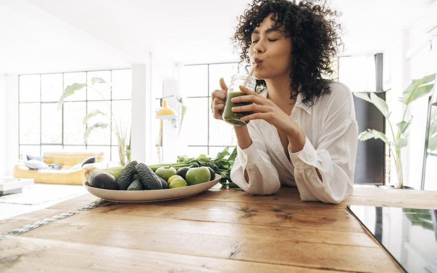 Woman in a white button up shirt sitting at a wooden kitchen table sipping a green smoothie out of a glass mug.