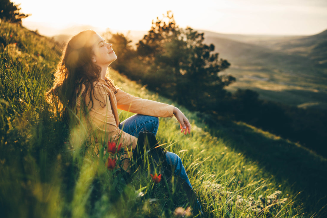 Young woman sitting in the grass on a hill with her eyes closed and her face tilted up toward the sky.