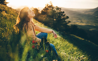 Young woman sitting in the grass on a hill with her eyes closed and her face tilted up toward the sky.