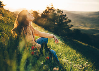 Young woman sitting in the grass on a hill with her eyes closed and her face tilted up toward the sky.