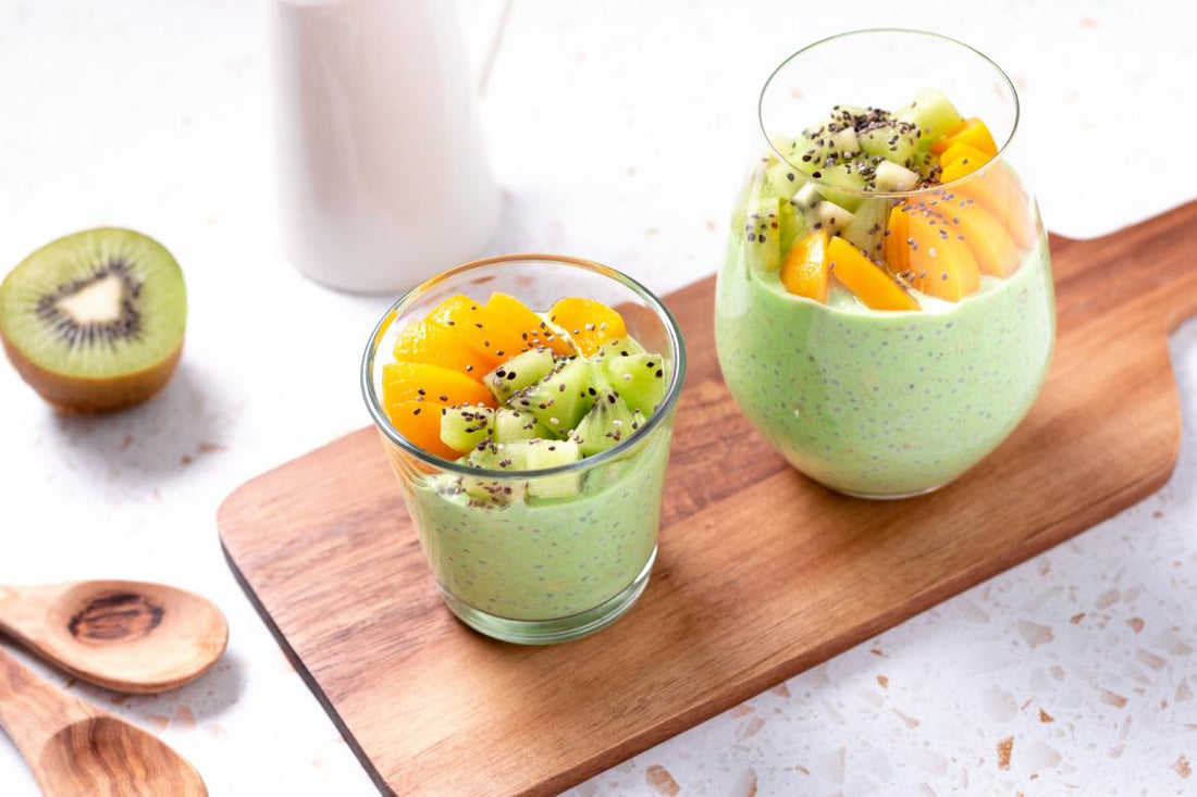 Two glasses filled with a green smoothie and topped with fruit sitting on a wooden cutting board on a kitchen countertop. 
