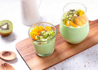 Two glasses filled with a green smoothie and topped with fruit sitting on a wooden cutting board on a kitchen countertop. 