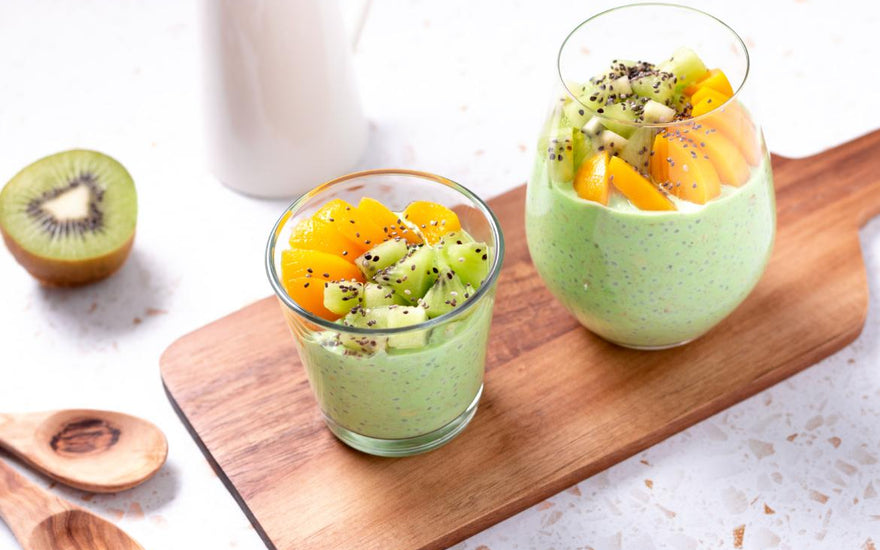 Two glasses filled with a green smoothie and topped with fruit sitting on a wooden cutting board on a kitchen countertop. 