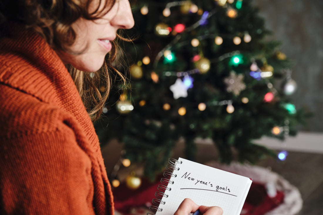 Smiling woman in an orange sweater sitting in front of a Christmas tree writing her New Year’s resolutions in a notebook. 