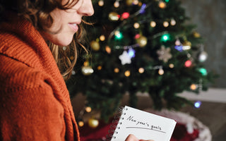 Smiling woman in an orange sweater sitting in front of a Christmas tree writing her New Year’s resolutions in a notebook. 