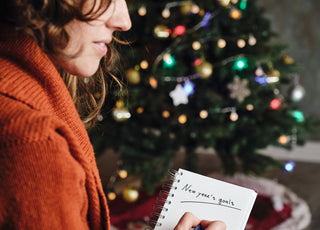 Smiling woman in an orange sweater sitting in front of a Christmas tree writing her New Year’s resolutions in a notebook. 