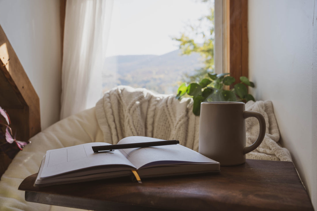 An open journal with a pen on top laying next to a brown mug on a side table placed next to a tan chair.