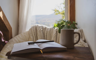 An open journal with a pen on top laying next to a brown mug on a side table placed next to a tan chair.