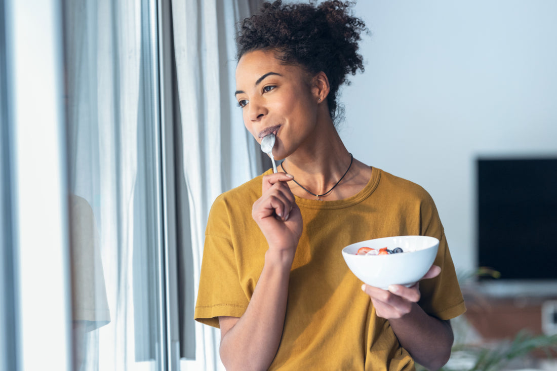 Woman in a yellow t-shirt holding a bowl of yogurt while looking out a window and pulling a metal spoon out of her mouth after taking a bite.