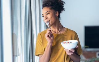 Woman in a yellow t-shirt holding a bowl of yogurt while looking out a window and pulling a metal spoon out of her mouth after taking a bite.