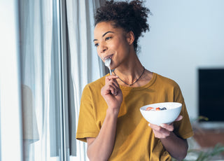 Woman in a yellow t-shirt holding a bowl of yogurt while looking out a window and pulling a metal spoon out of her mouth after taking a bite.