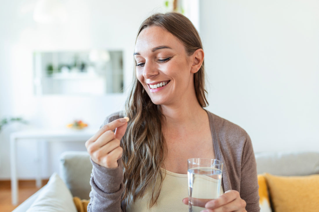 Woman sitting on a couch with a glass of water in one hand while she happily examines a small pill she is holding in the other hand.