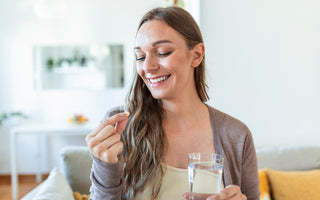 Woman sitting on a couch with a glass of water in one hand while she happily examines a small pill she is holding in the other hand.