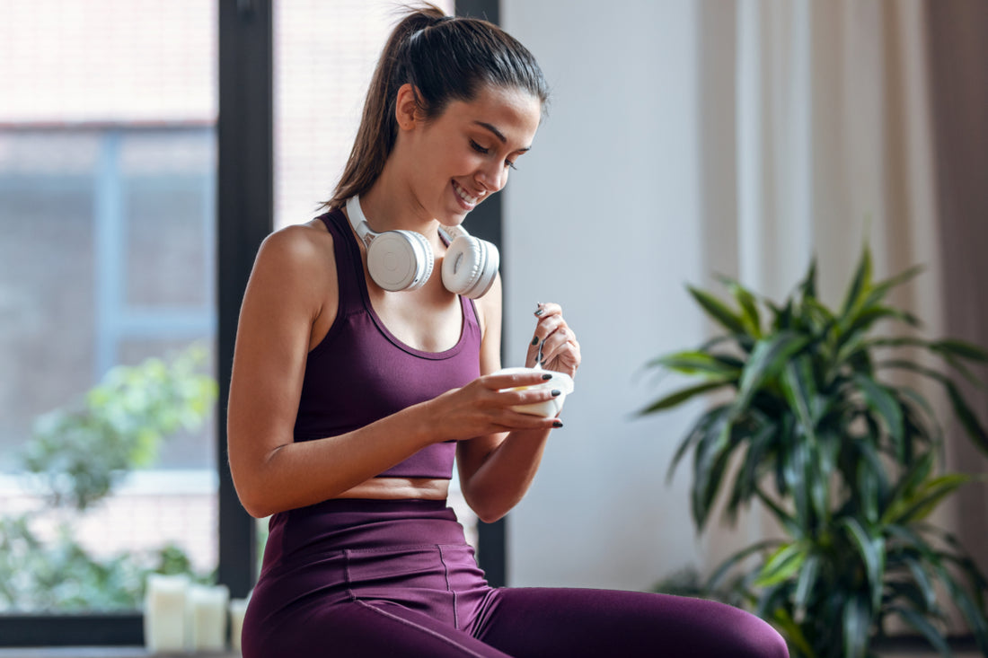 Woman in maroon workout clothes and white headphones around her neck sitting and enjoying a cup of yogurt.