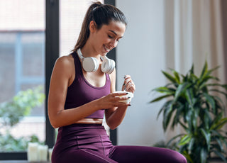 Woman in maroon workout clothes and white headphones around her neck sitting and enjoying a cup of yogurt.