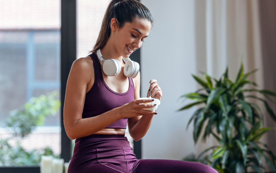 Woman in maroon workout clothes and white headphones around her neck sitting and enjoying a cup of yogurt.