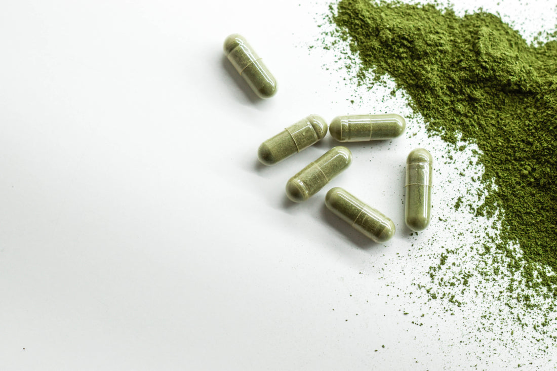 Six green supplement capsules laying on a white surface next to a pile of green powder.