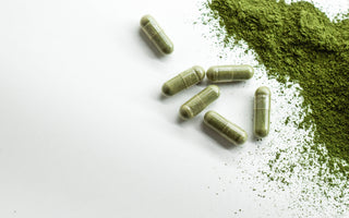 Six green supplement capsules laying on a white surface next to a pile of green powder.