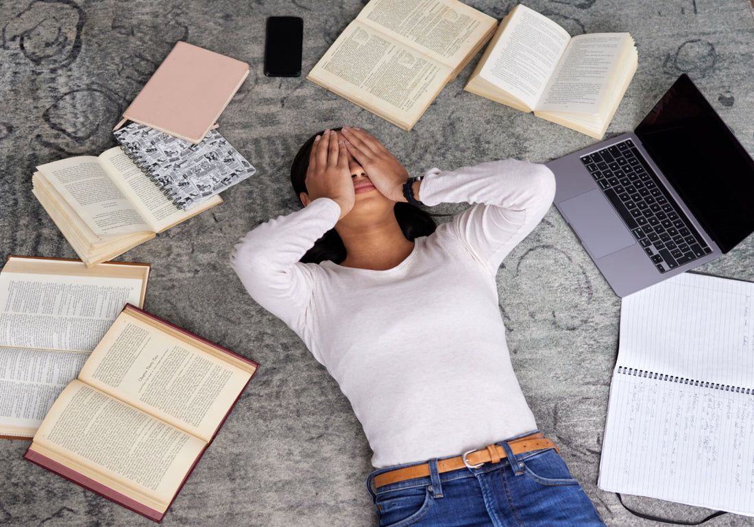 Young adult women laying on the floor surrounded by books and notebooks. She has her hands over her eyes as if stressed.