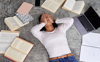 Young adult women laying on the floor surrounded by books and notebooks. She has her hands over her eyes as if stressed.