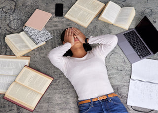 Young adult women laying on the floor surrounded by books and notebooks. She has her hands over her eyes as if stressed.