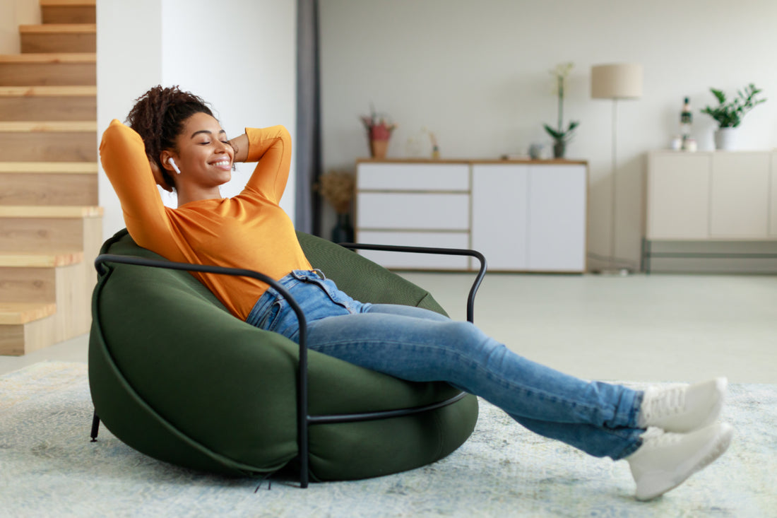 Woman in an orange shirt and jeans reclining on a bean bag chair with her eyes closed while listening to something via wireless ear buds.