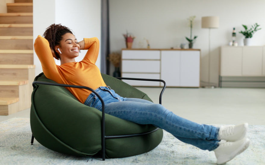 Woman in an orange shirt and jeans reclining on a bean bag chair with her eyes closed while listening to something via wireless ear buds.