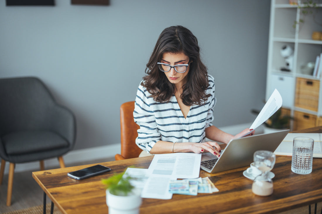 Business woman wearing a black and white striped top and glass sitting at a wooden desk examining a printed report while working on her laptop.