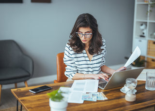 Business woman wearing a black and white striped top and glass sitting at a wooden desk examining a printed report while working on her laptop.