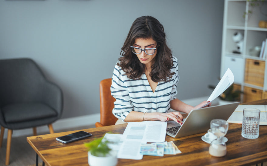 Business woman wearing a black and white striped top and glass sitting at a wooden desk examining a printed report while working on her laptop.