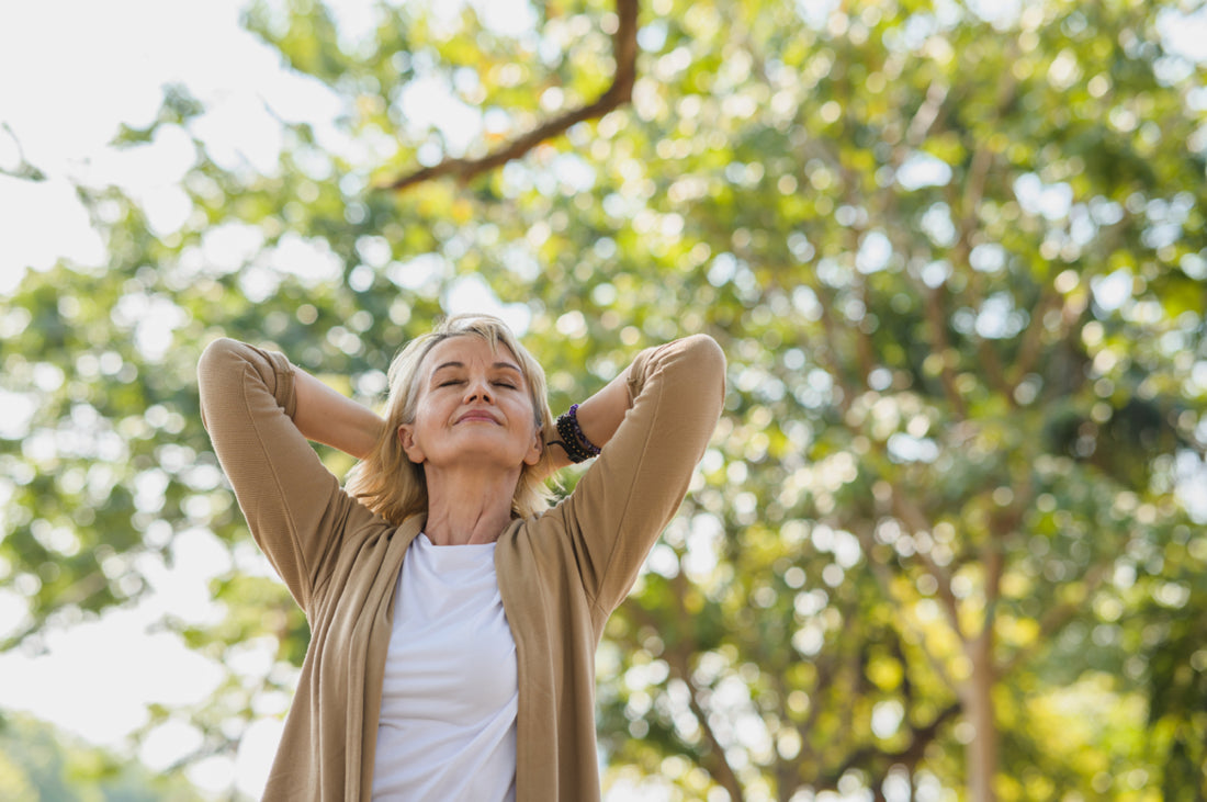 Middle-aged woman standing outside under a tree with her hands behind her head, her face tilted up, and her eyes closed.
